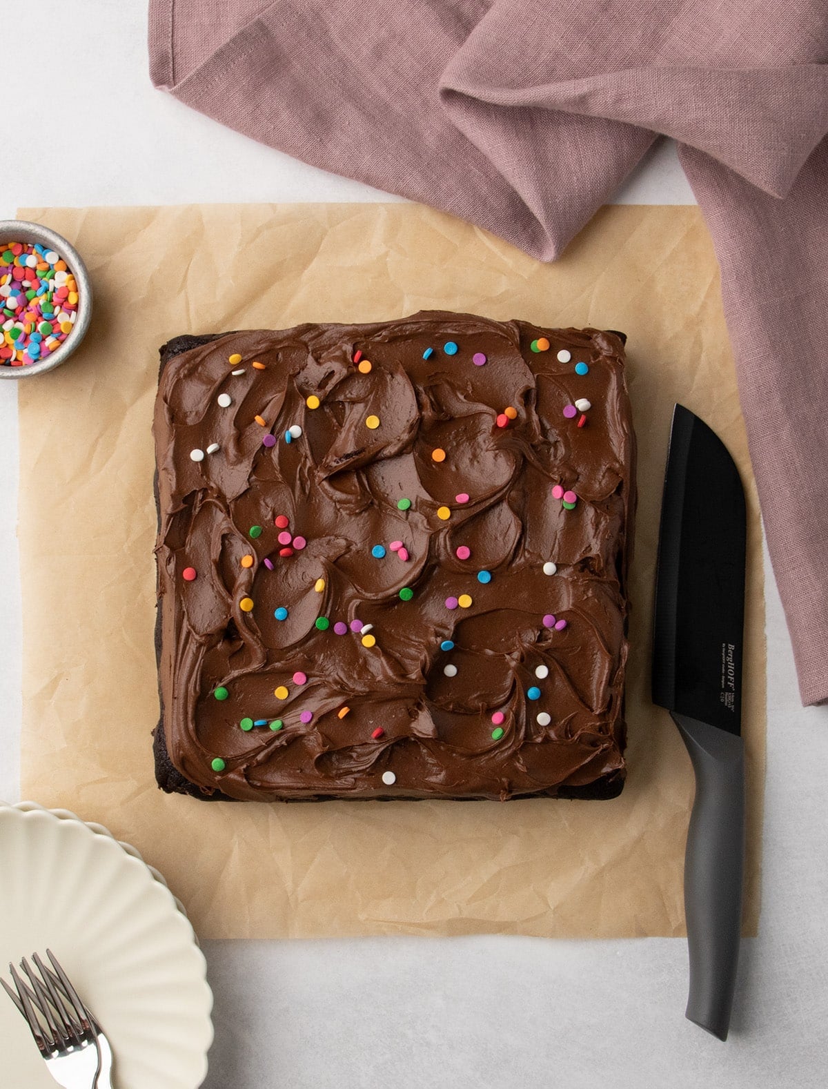Overhead view of a frosted square chocolate cake with colorful sprinkles on parchment paper, with a knife and a small bowl of sprinkles nearby.