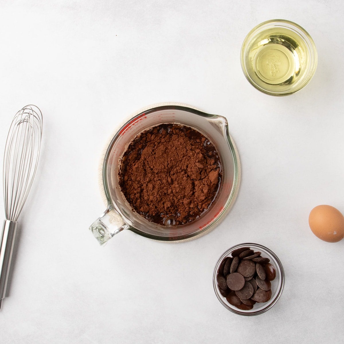 Cocoa powder sitting on top of liquid in a glass measuring cup, with a whisk, a small bowl of chocolate discs, a whole egg, and a small bowl of oil nearby.