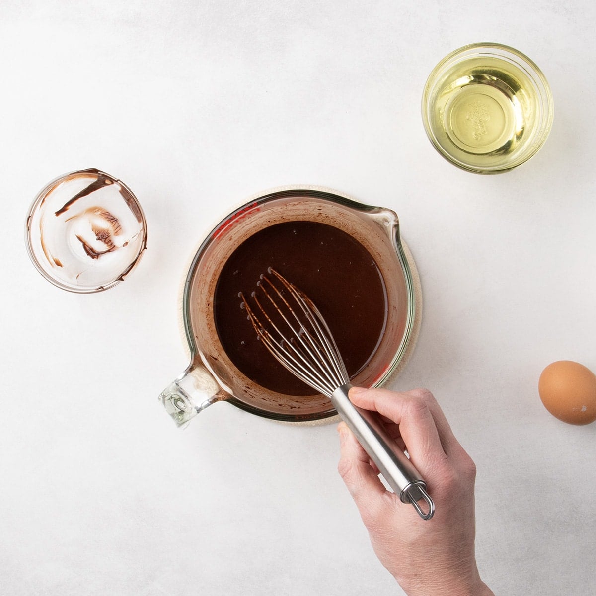 A hand holding a whisk mixing a dark chocolate liquid in a glass measuring cup, with a small empty bowl, a whole egg, and a small bowl of oil nearby.