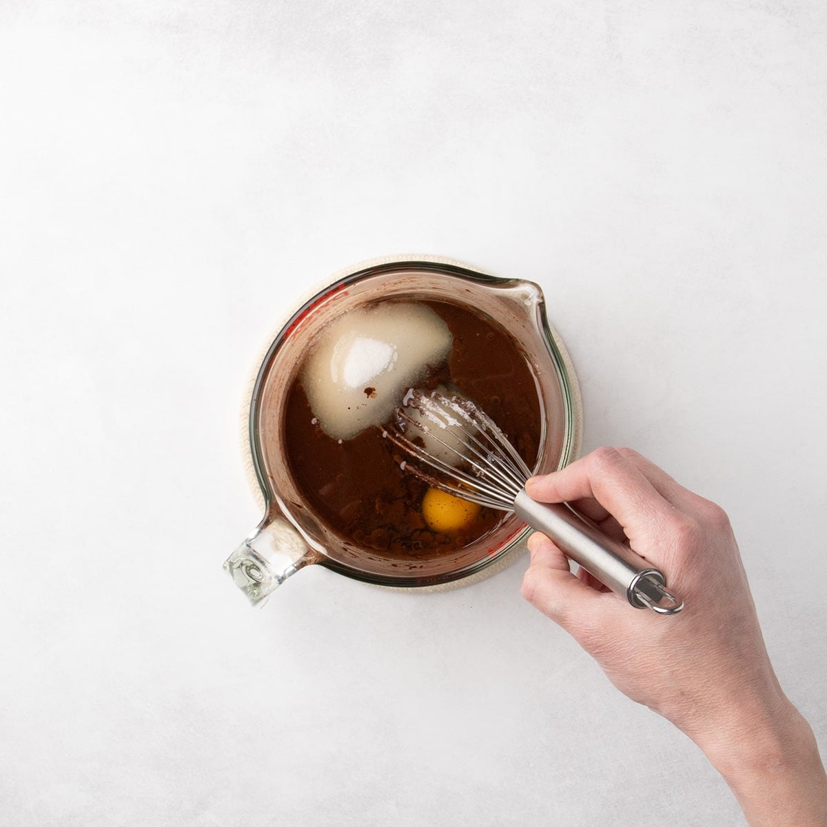 A hand whisking a chocolate mixture in a glass measuring cup with a whole egg and granulated sugar soaking into the liquid.