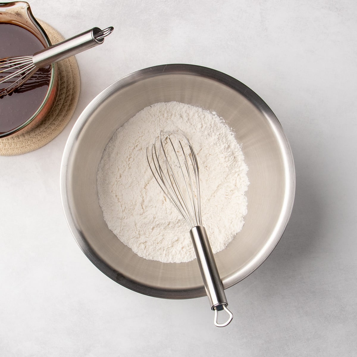 A metal mixing bowl filled with a white flour mixture and a whisk resting inside it, with a glass measuring cup of chocolate mixture partially visible.