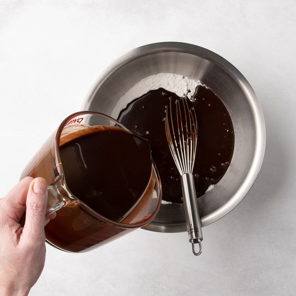Chocolate mixture being poured from a glass measuring cup into a metal bowl with a white flour mixture with a whisk inside.