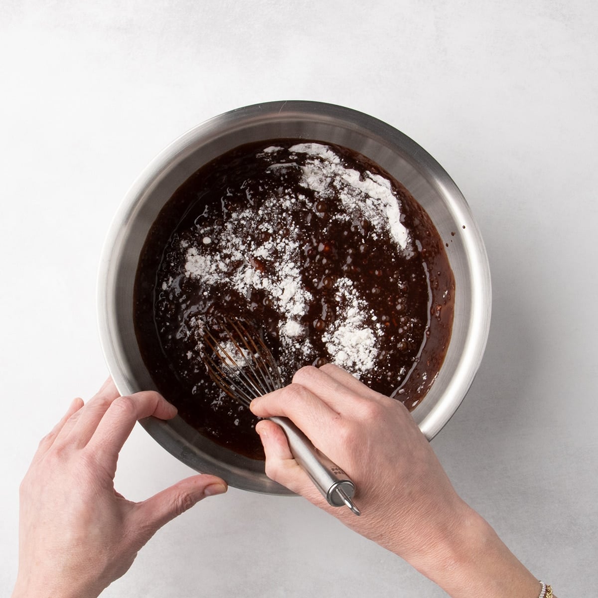 Hands holding a whisk and mixing a dark batter with patches of white flour still visible in a metal bowl.