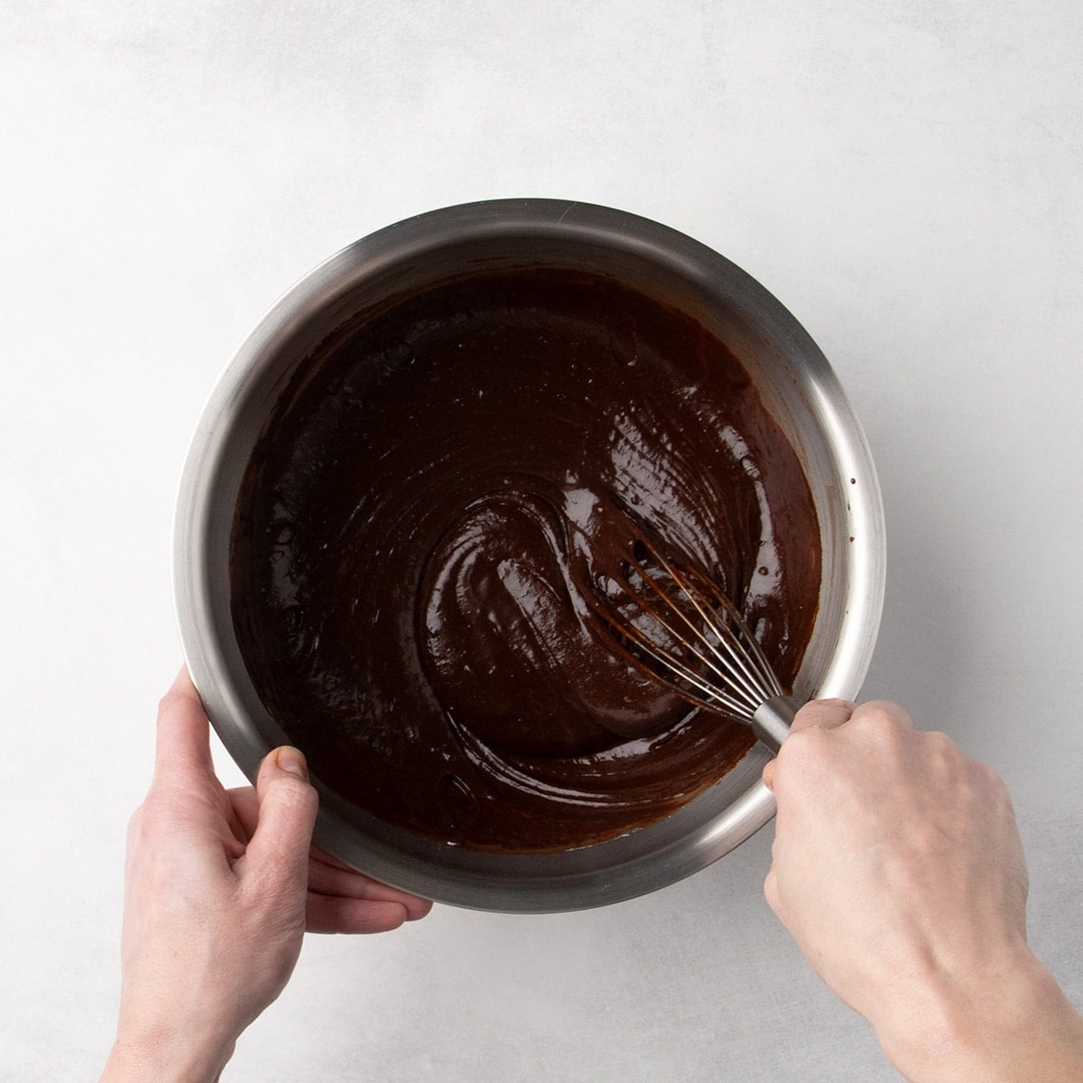 A hand using a whisk to mix a thick, smooth dark chocolate batter in a metal bowl.