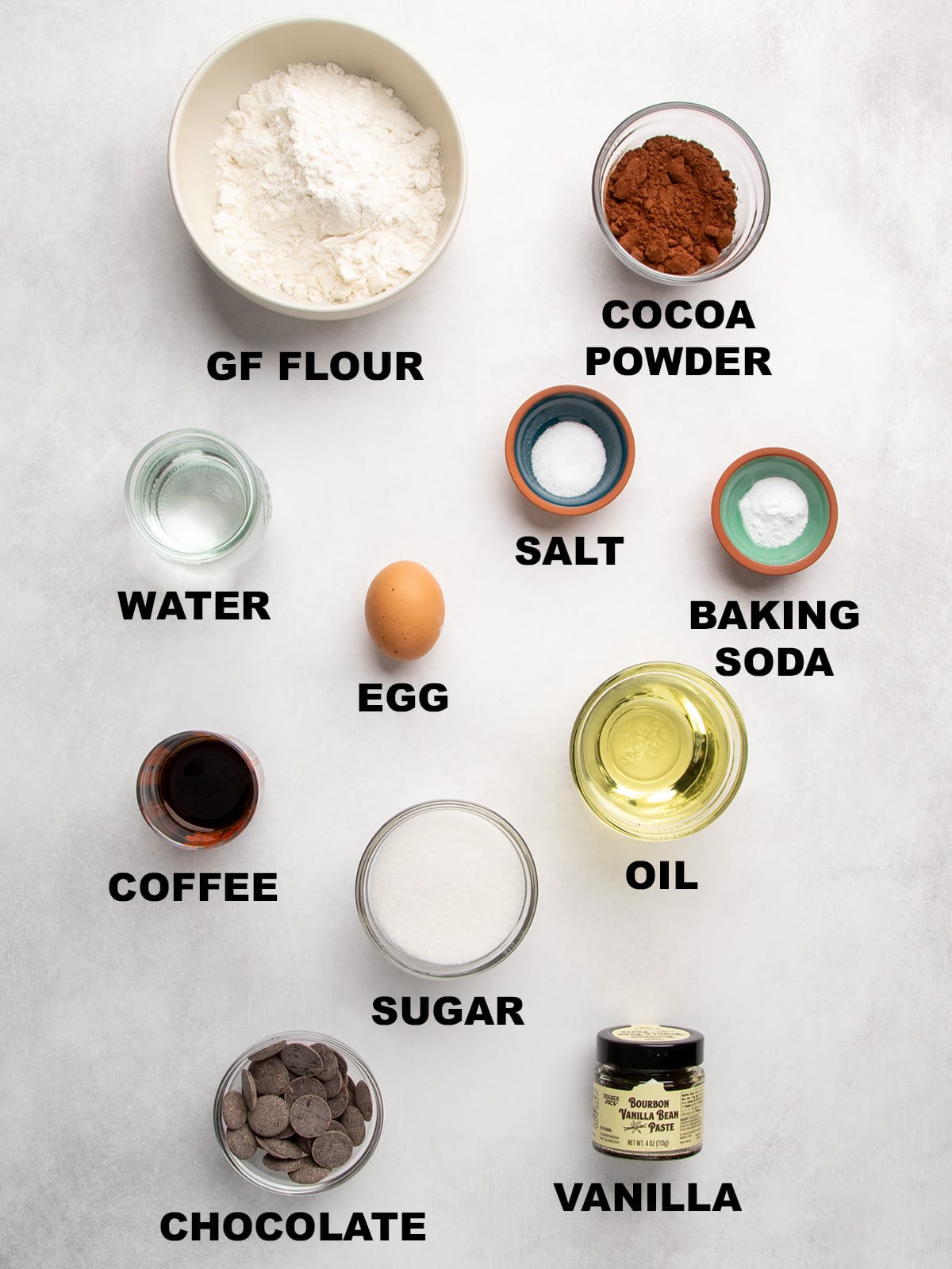 Overhead view of labeled ingredients including gluten free flour, cocoa powder, salt, baking soda, water, egg, coffee, oil, sugar, chocolate, and vanilla bean paste in a jar.