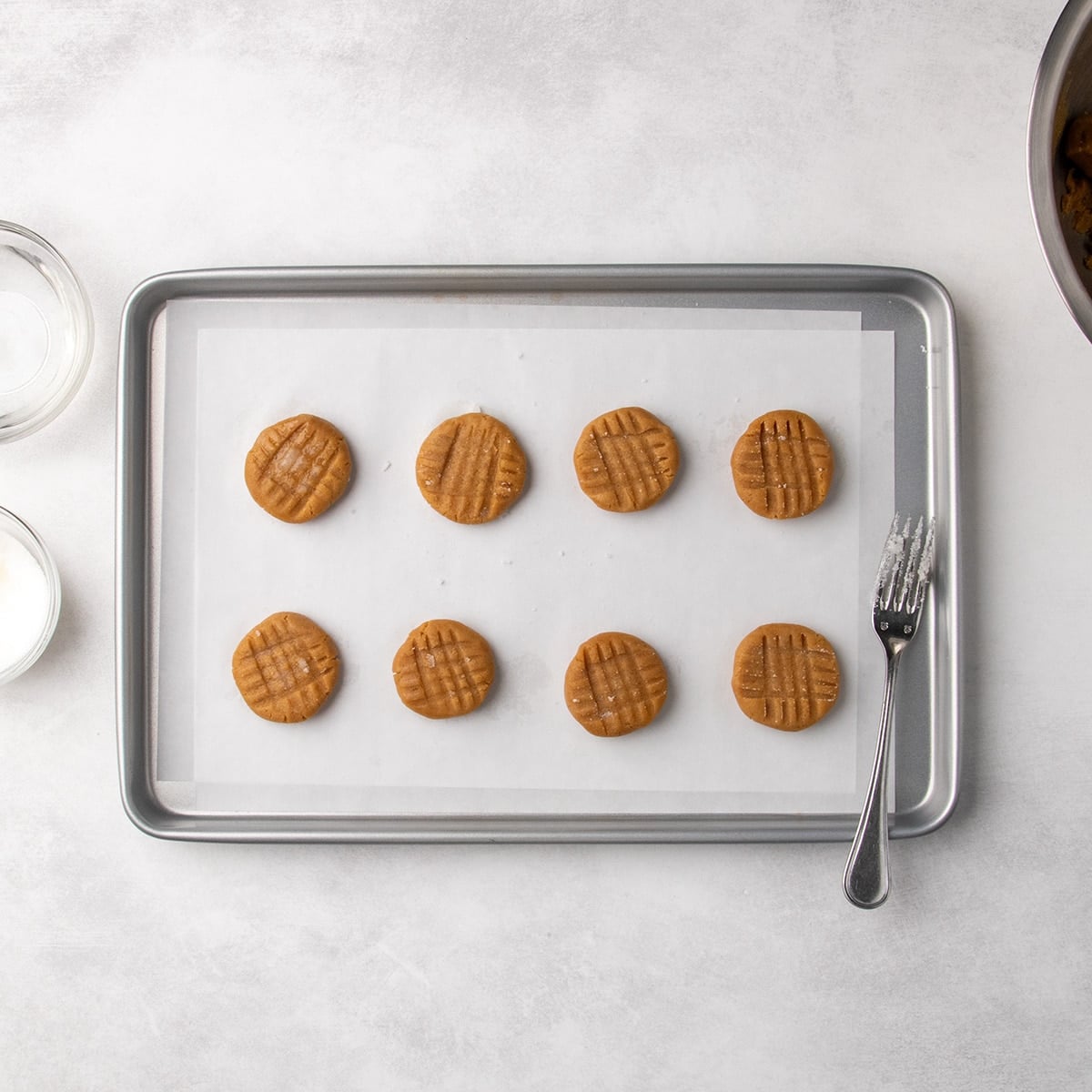Flattened peanut butter cookie dough rounds with fork crosshatch marks arranged in rows on a parchment-lined baking sheet with a fork on the side.
