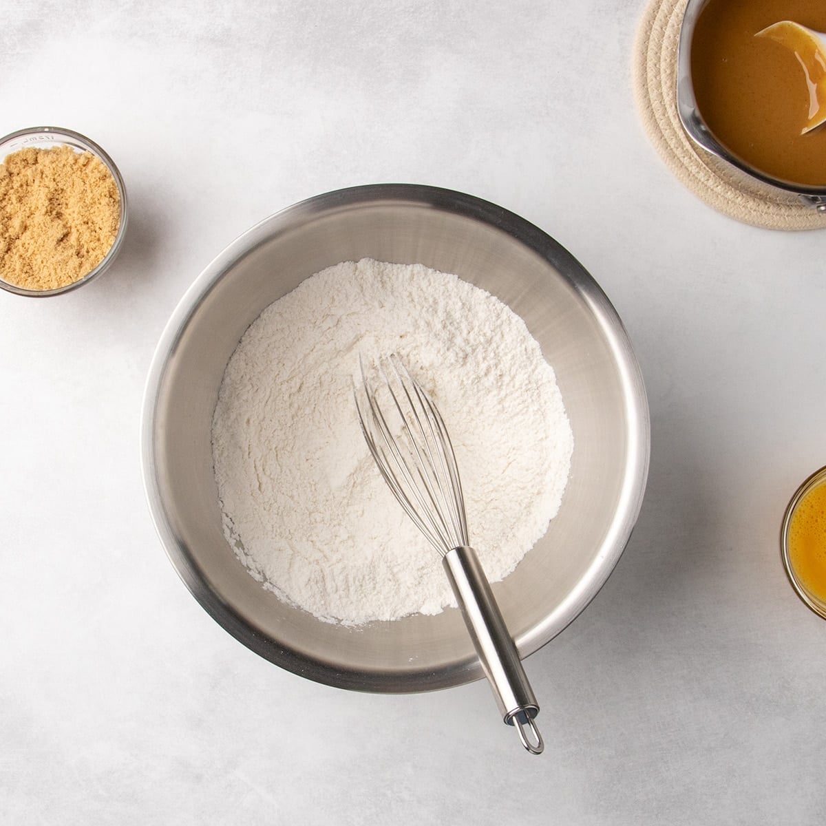 Large metal mixing bowl filled with white dry ingredients and a whisk, with a small bowl of brown sugar and a saucepan of peanut butter mixture nearby.