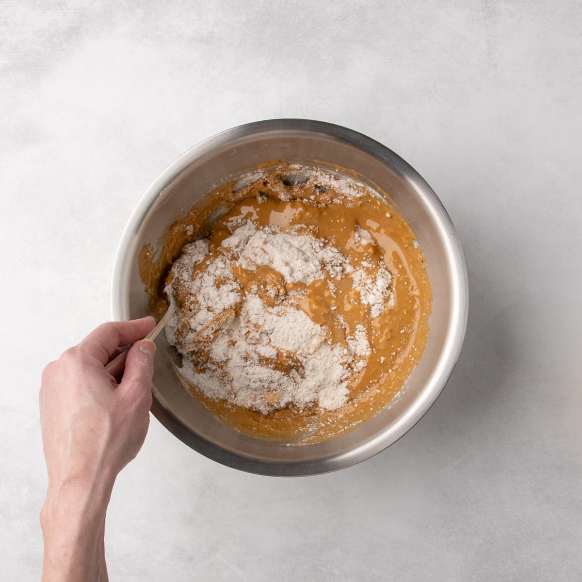 Hand stirring peanut butter mixture and flour mixture together in a metal mixing bowl with a spoon.