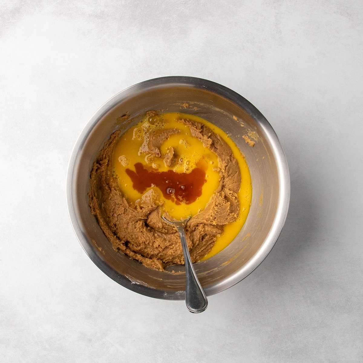Egg mixture and vanilla added on top of peanut butter dough in a metal mixing bowl with a spoon.