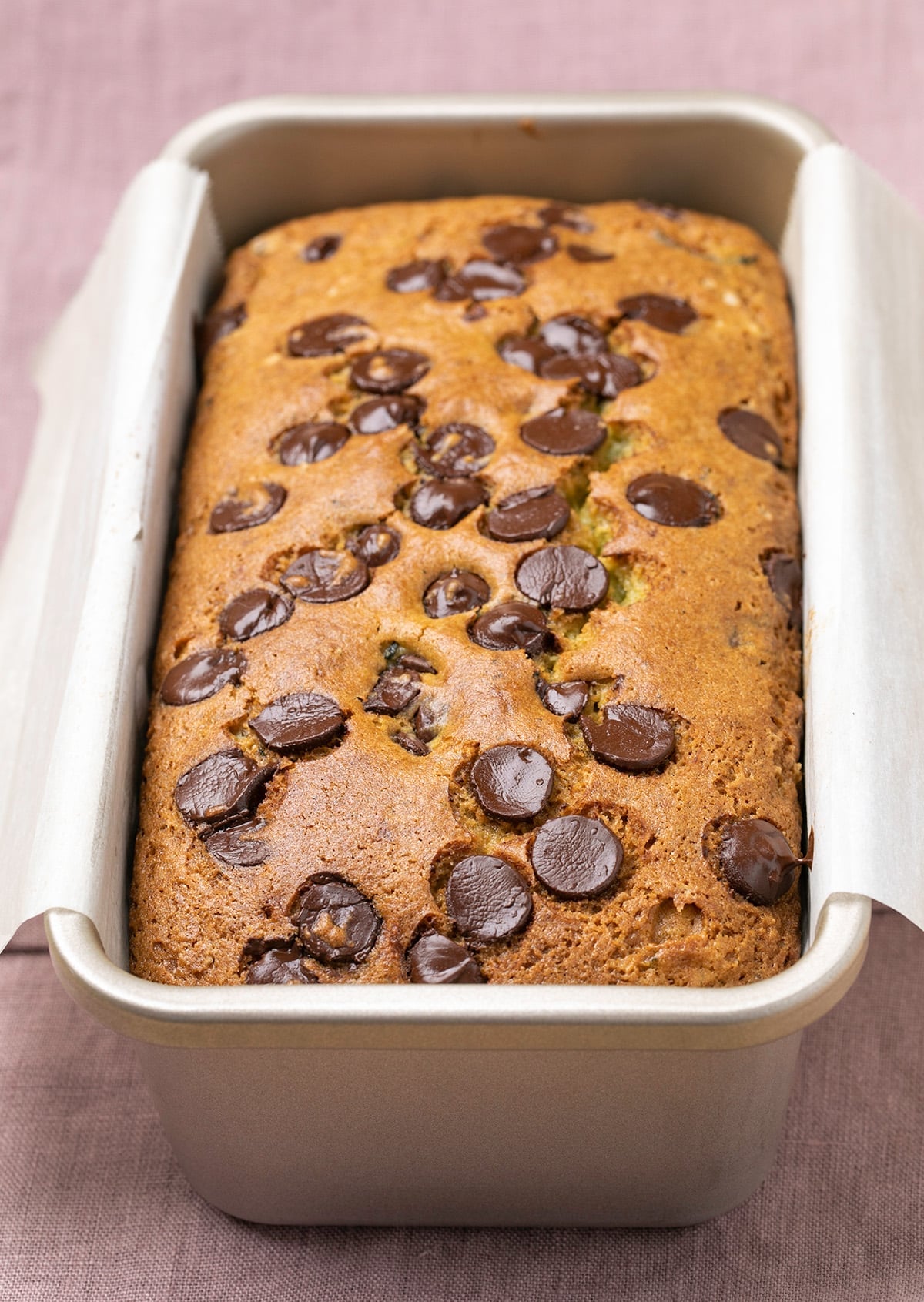 The bread with brown spots in nonstick loaf pan lined with white paper on purple cloth.