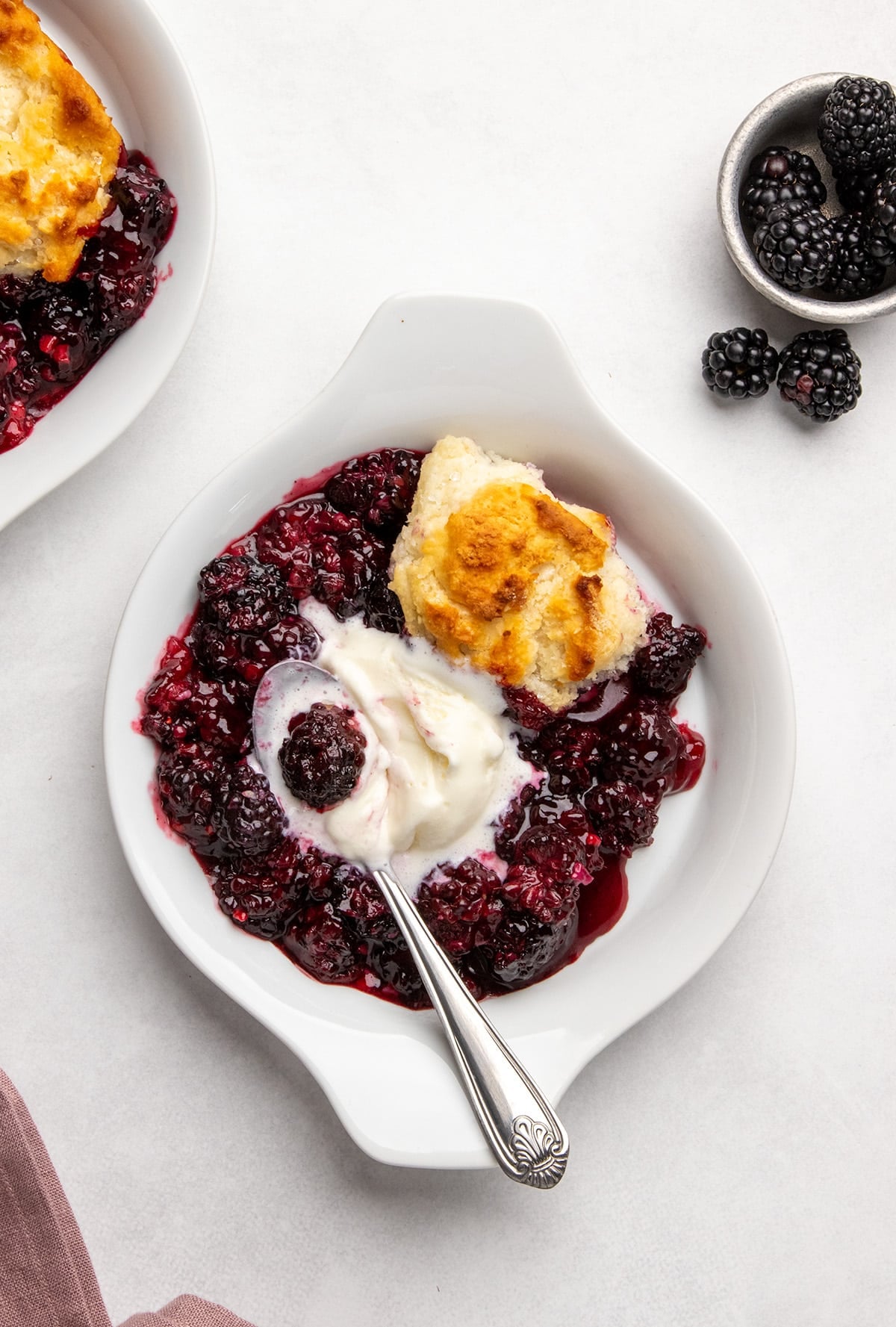 small round dish with blackberry cobbler, melting vanilla ice cream, a spoon, and fresh blackberries nearby