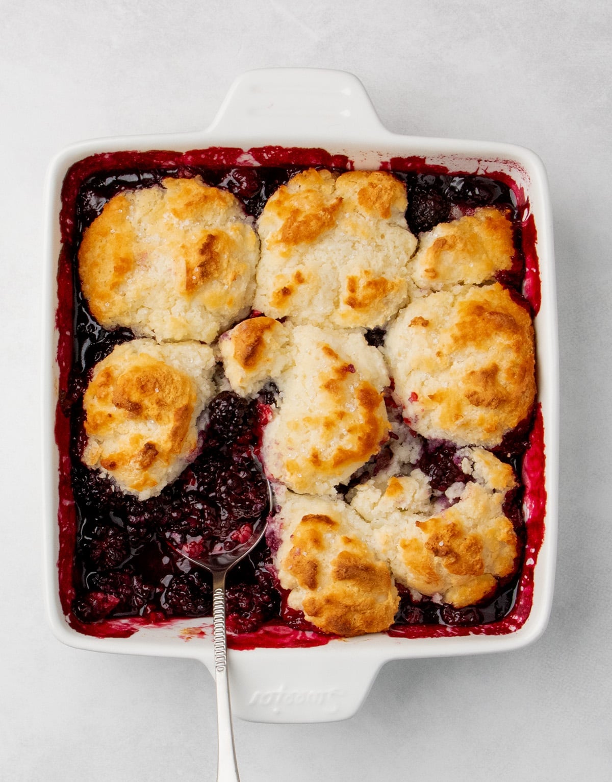 overhead view of baked blackberry cobbler in a square white baking dish with a spoon in the berry filling