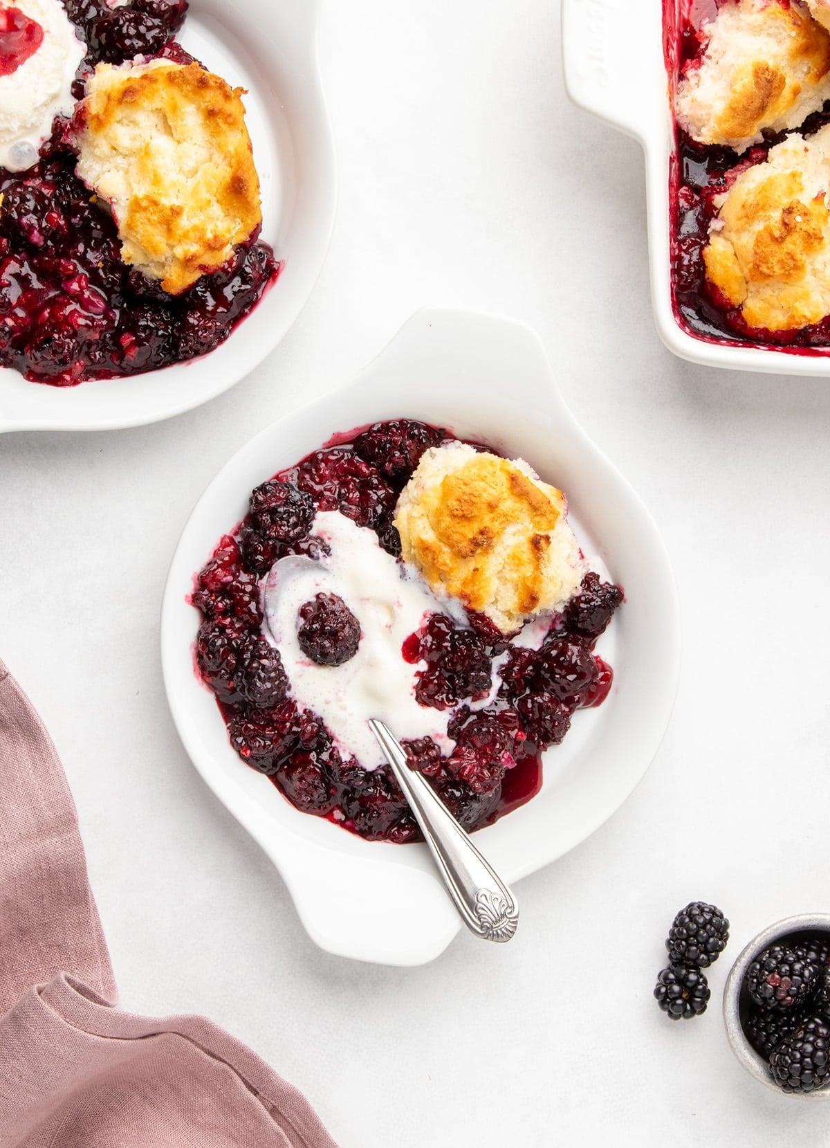 serving dish of blackberry cobbler with mostly melted vanilla ice cream and a spoon, with more cobbler and fresh blackberries nearby