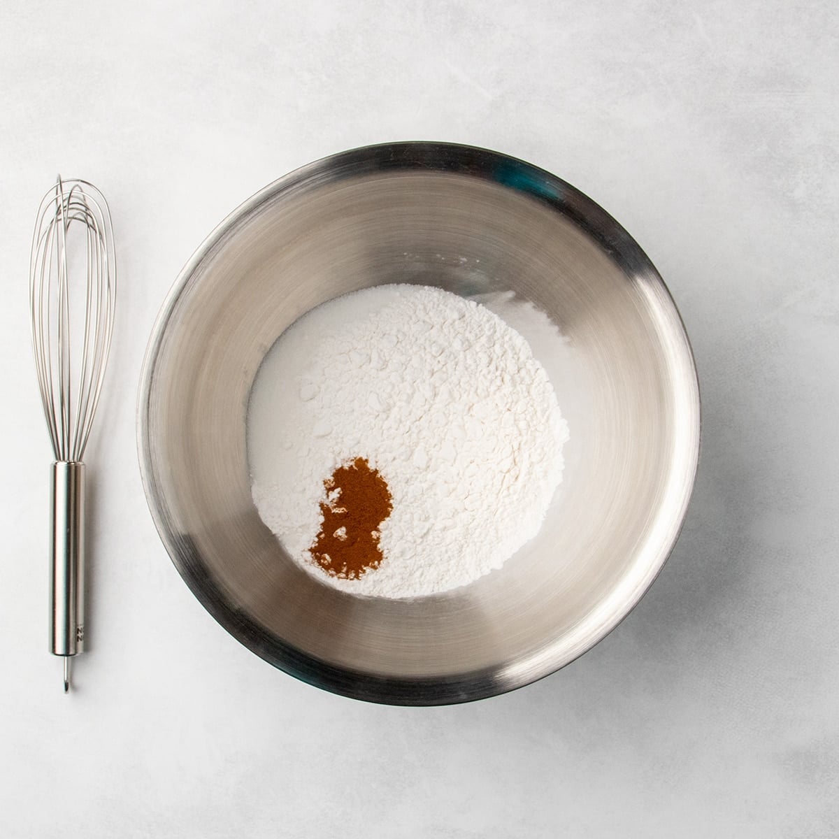 metal mixing bowl with white dry ingredients and a small pile of brown dried cinnamon spice, whisk placed beside the bowl