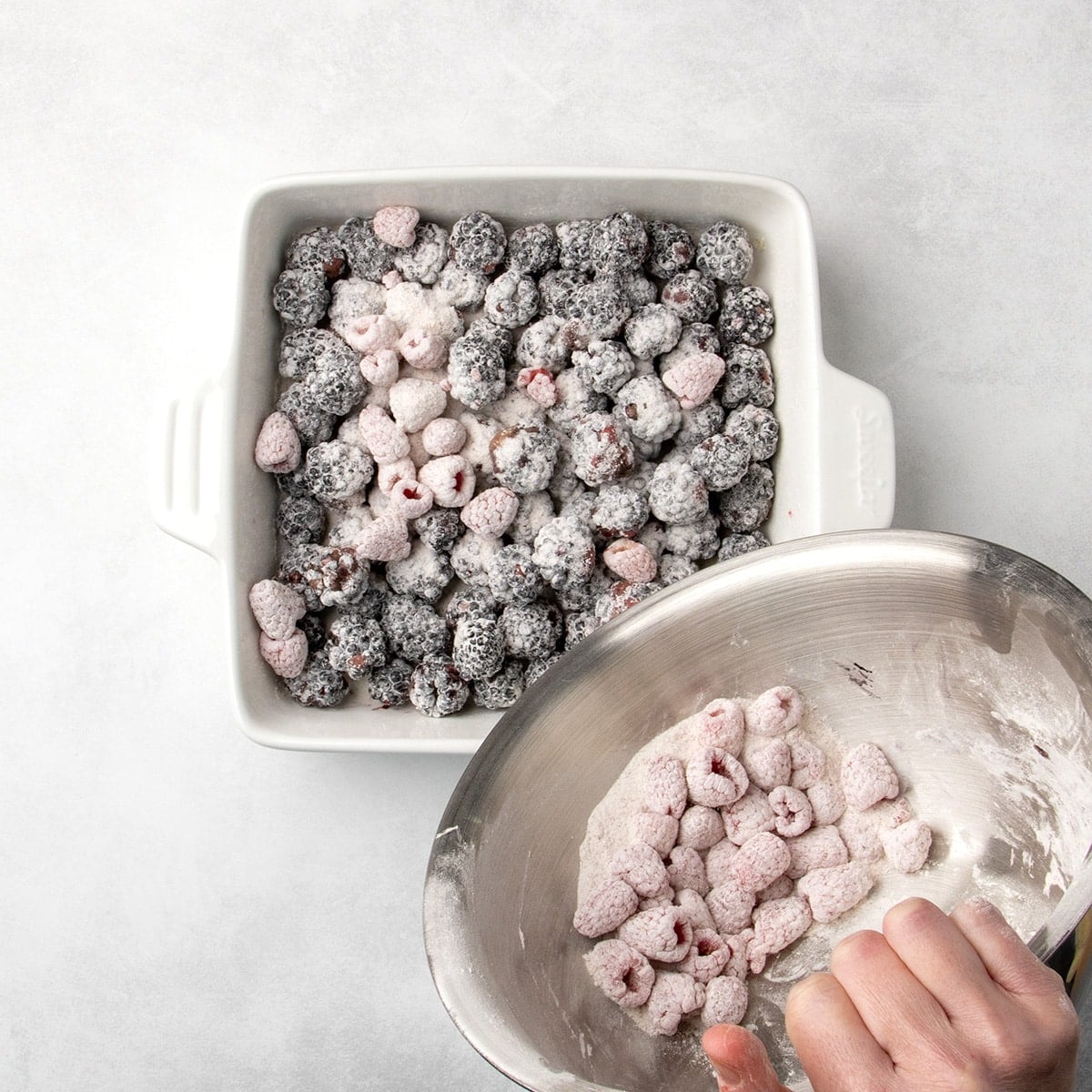 red raspberries coated in white powder being transferred from a metal bowl into a white baking dish with blackberries coated in the same powder