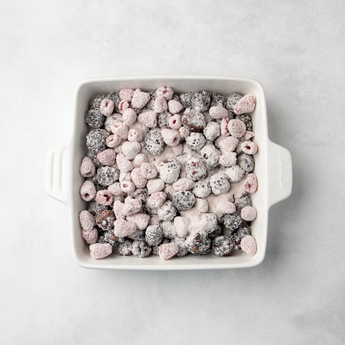 blackberries and raspberries coated in white powder spread in an even layer in a square white baking dish