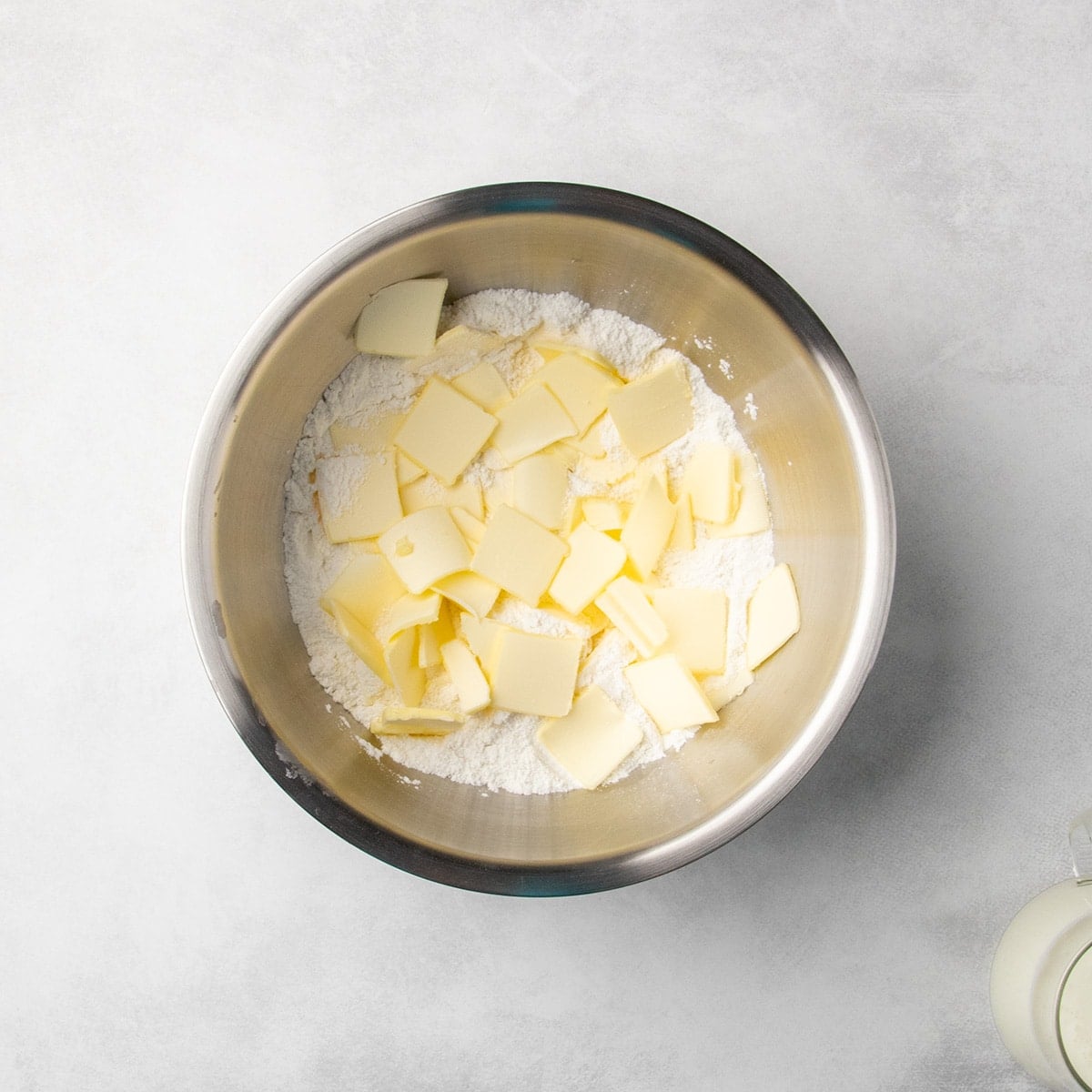 metal mixing bowl with white dry ingredients topped with small, mostly square pieces of sliced butter