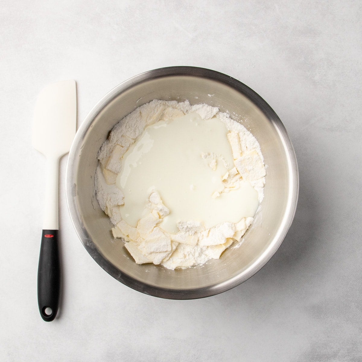 metal bowl with dry ingredients, chunks of butter, and white liquid buttermilk added, spatula placed beside the bowl