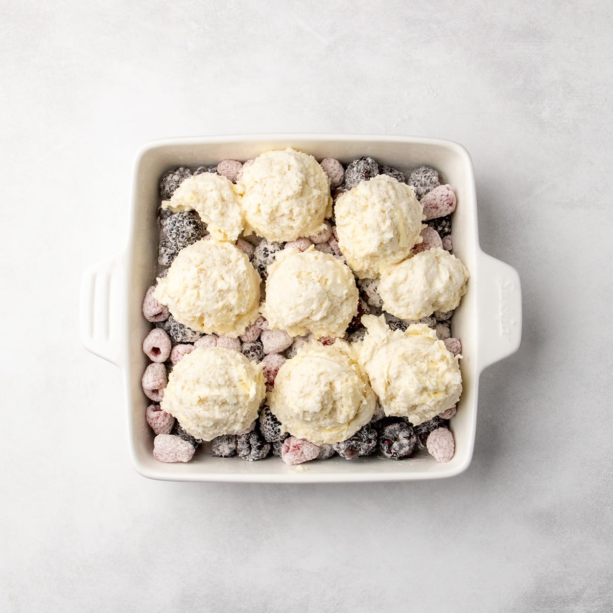 scooped mounds of pale raw dough mixture placed on top of the berry and flour mixture in a white baking dish