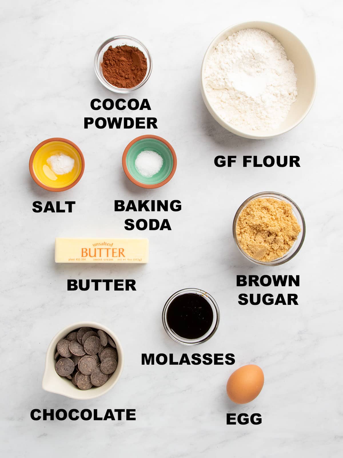 Bowls of cocoa powder, gluten free flour, salt, baking soda, brown sugar, chocolate pieces, molasses, butter, and an egg arranged on a marble surface with labels.