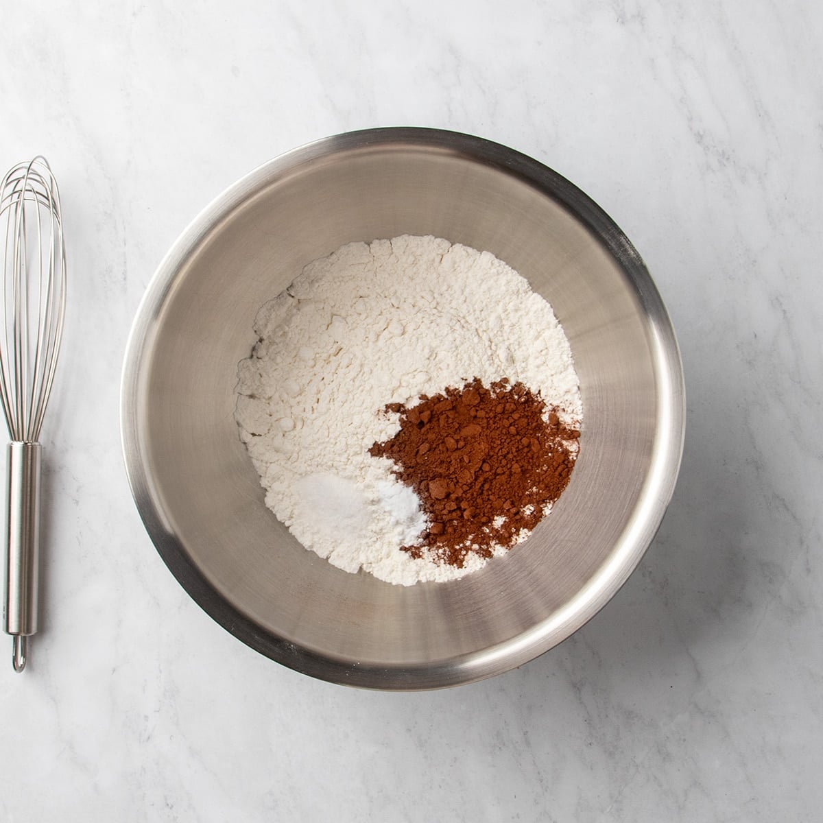 Metal mixing bowl with white powder and cocoa powder, with a whisk beside the bowl on a marble surface.