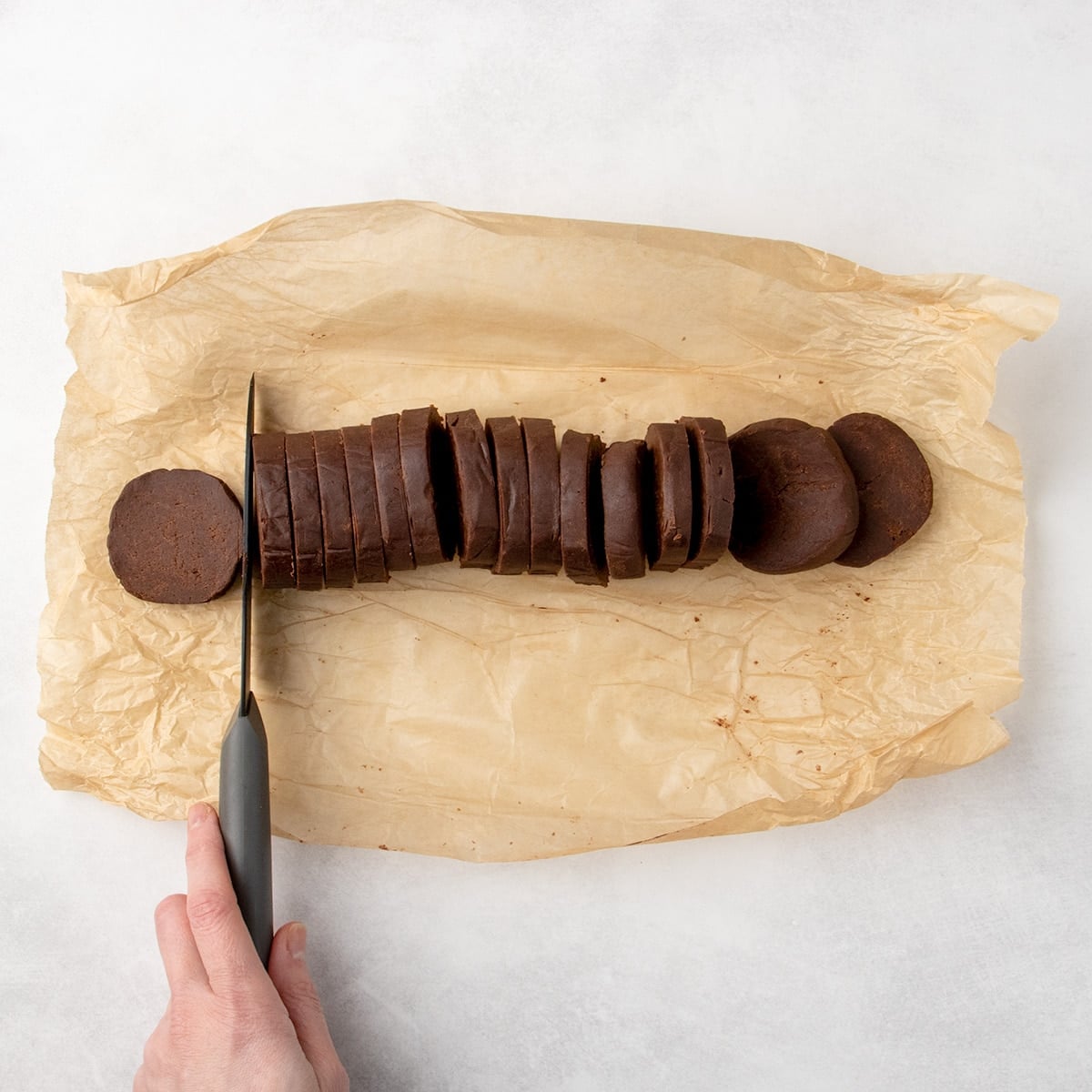 Sliced chocolate dough log on parchment paper with a hand holding a black knife cutting more slices into the remaining whole, still on brown parchment paper.