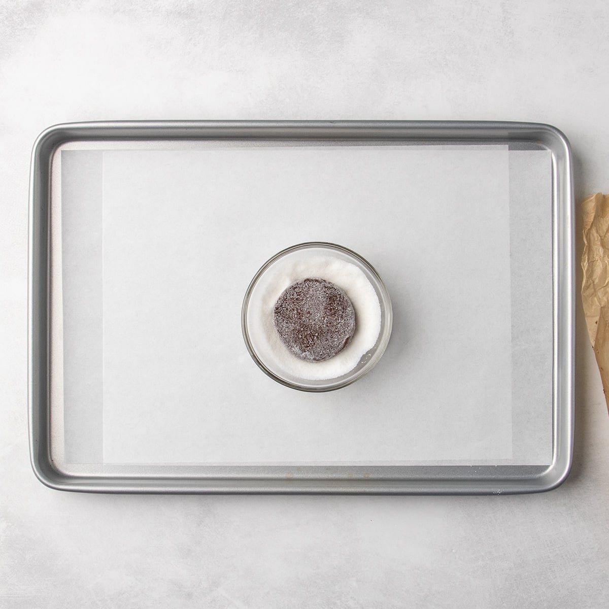 Round piece of chocolate dough coated in white sugar in a small glass bowl on a parchment-lined baking sheet.