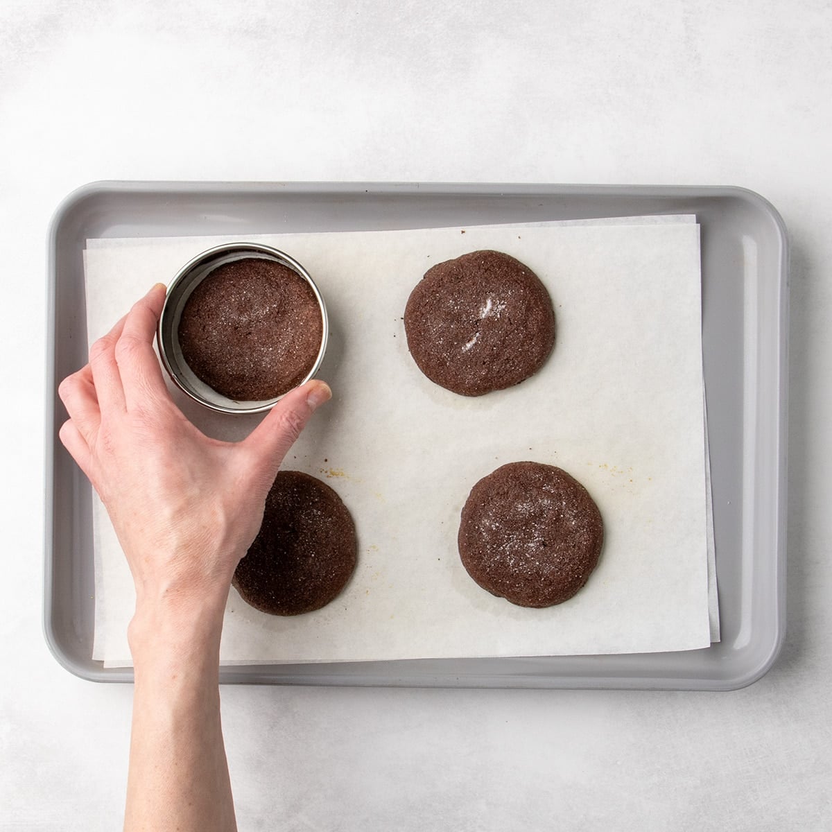 Hand placing a round cutter over a baked chocolate cookie on a parchment-lined baking sheet, coaxing it into a proper round while still hot from the oven.
