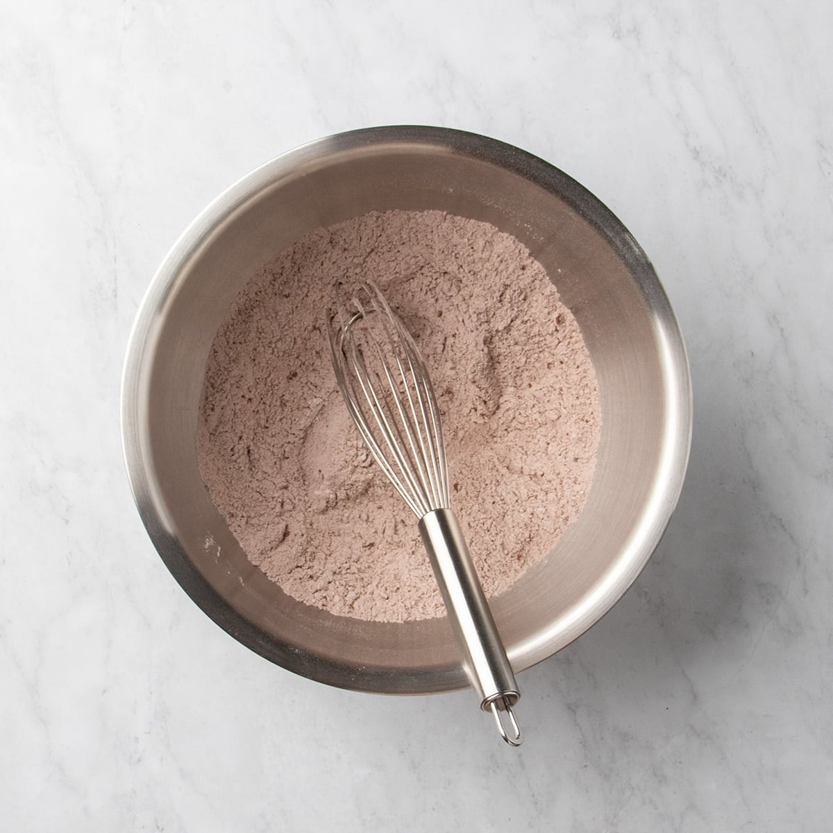 Metal mixing bowl with light brown dry mixture and a whisk resting inside on a marble surface.