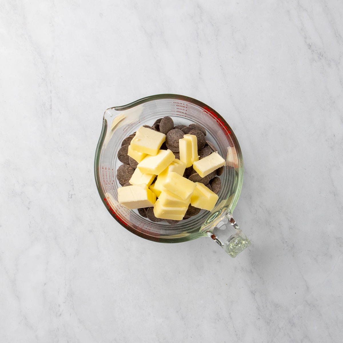 Glass measuring cup filled with chocolate pieces and small cubes of butter, all resting on a marble surface.