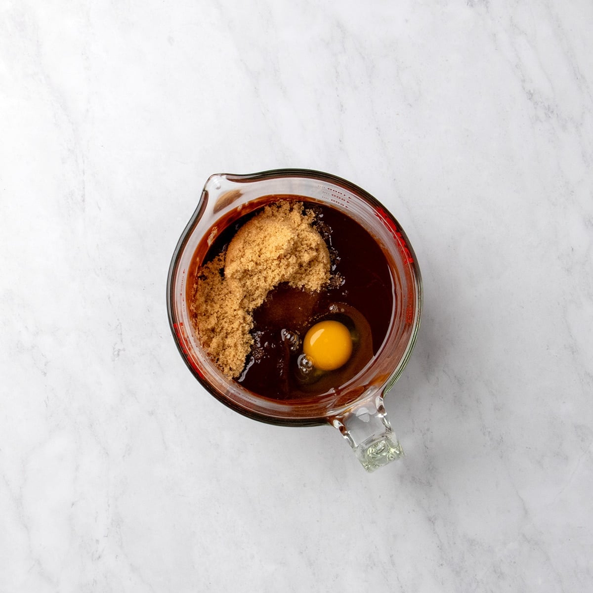 Same glass measuring cup now with melted chocolate mixture, and brown sugar, and a whole raw unbeaten egg sitting on top.