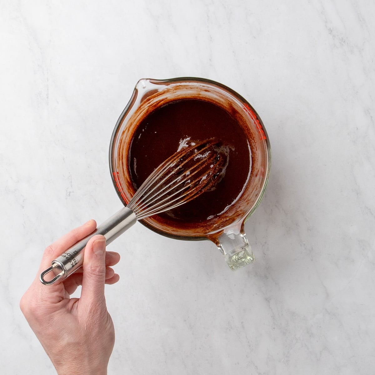 Hand holding a whisk mixing a dark chocolate mixture in a glass measuring cup.