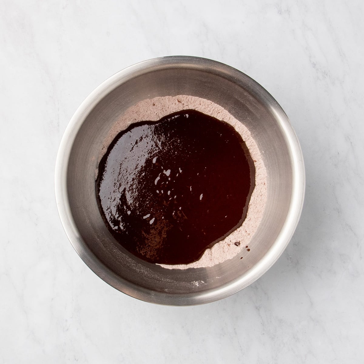 Melted chocolate mixture unmixed on top of pale brown dry ingredients in a metal mixing bowl.