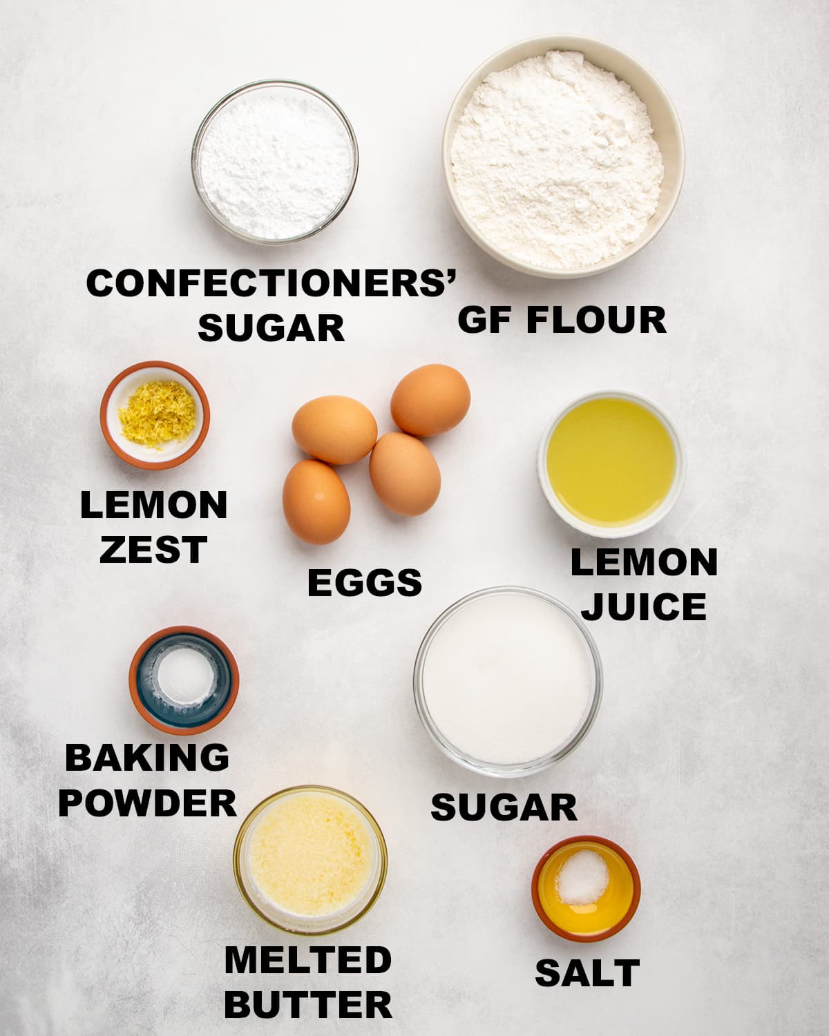 Overhead view of measured ingredients in bowls with labels including confectioners’ sugar, gluten free flour, lemon zest, eggs, lemon juice, baking powder, sugar, melted butter, and salt.