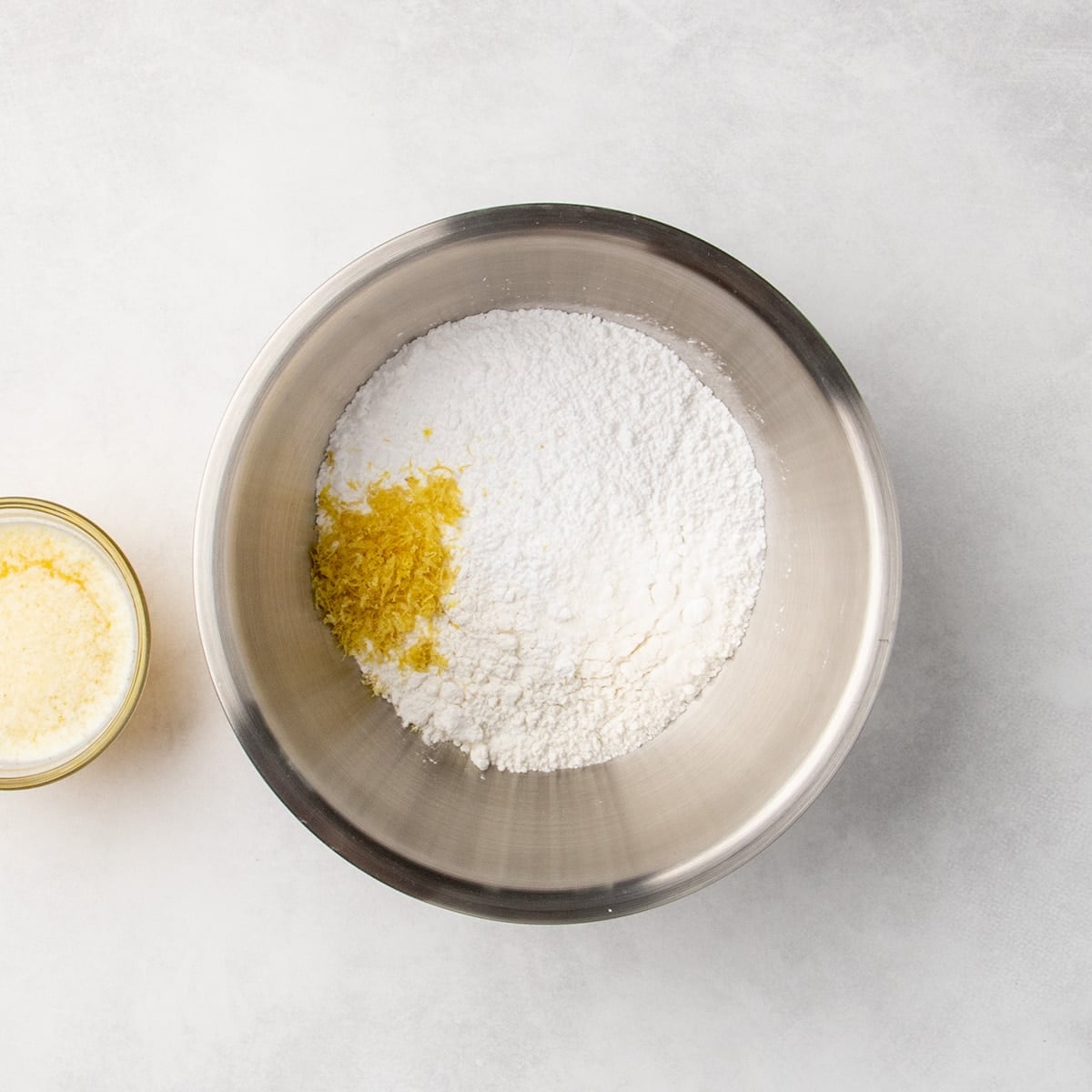 Metal mixing bowl filled with white dry ingredients and a small pile of yellow lemon zest, with a glass of melted butter mixture partially visible to the side.