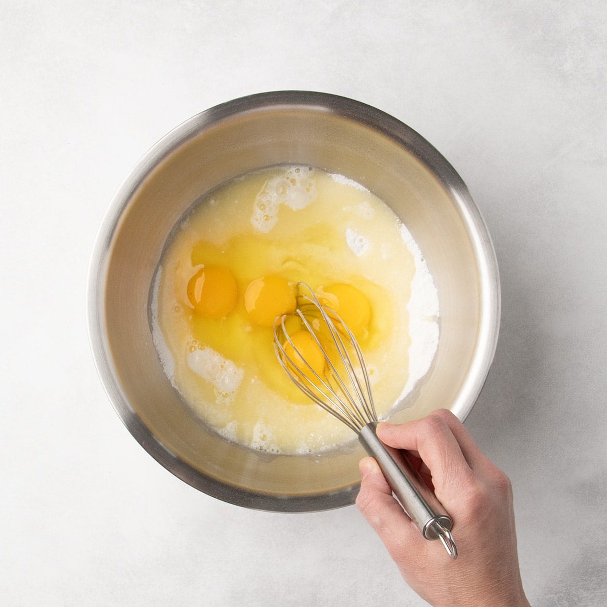 Hand holding a metal whisk about to whisk together a mixture of white dry ingredients with eggs and lemon juice liquid mixture.