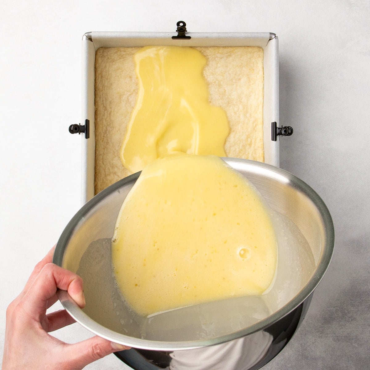 Hand pouring a pale yellow liquid filling mixture from a metal bowl into a baked crust in a parchment-lined square pan.