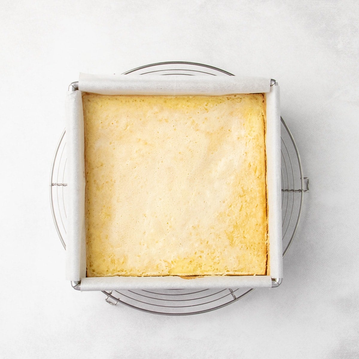 Uncut, baked lemon bars in a parchment-lined square pan resting on a round wire rack, lightly golden around the edges.