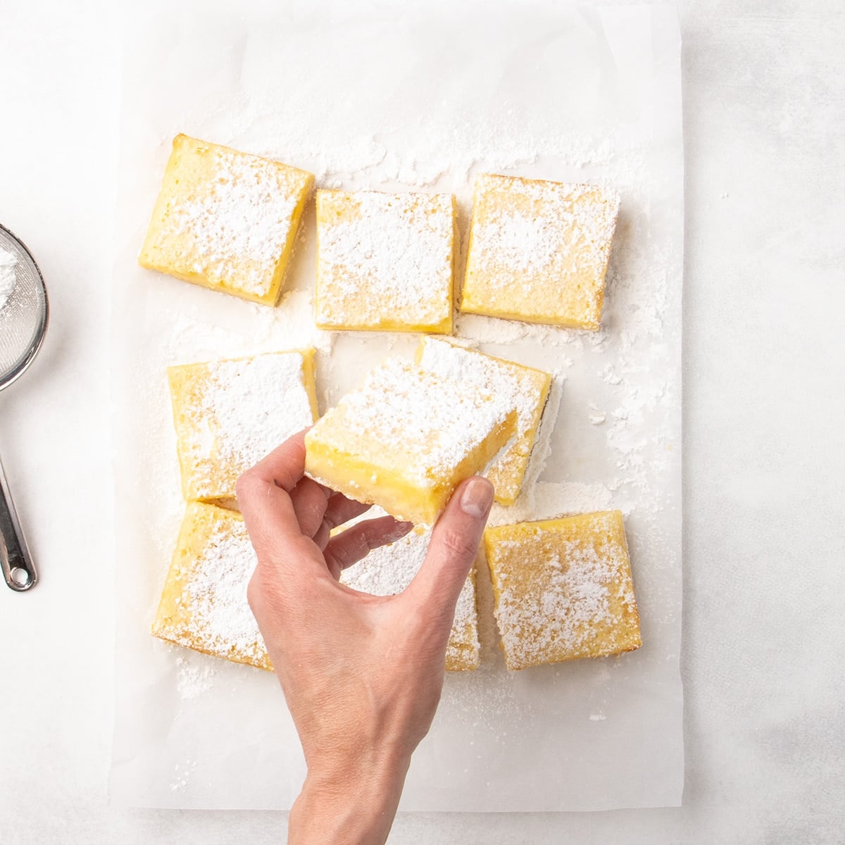 Hand holding a square lemon bar dusted with powdered sugar over several cut bars on parchment paper, with a sieve partially visible to the side.