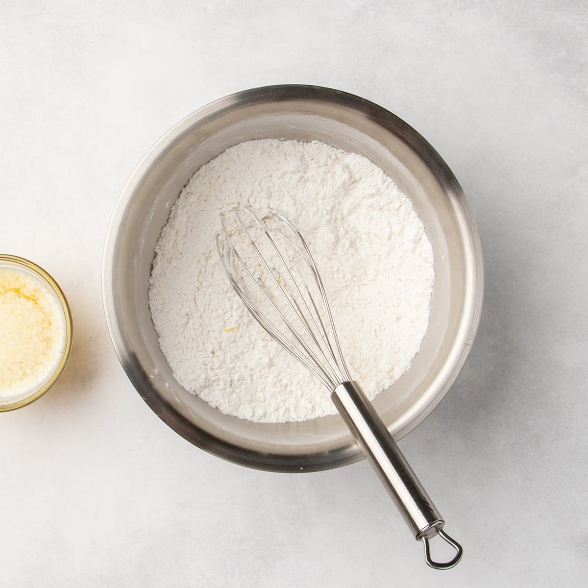 Metal mixing bowl with white dry ingredients being whisked with a metal whisk, with some visible yellow flecks of lemon zest