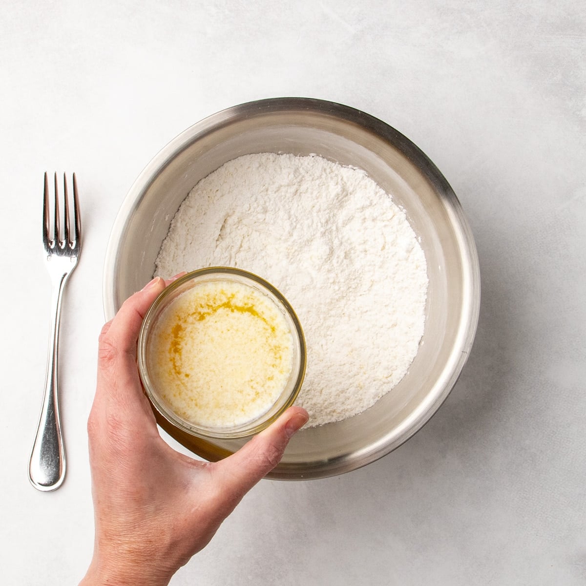 Hand holding a small glass of melted butter mixture about to pour it into a bowl of white dry ingredients, with a fork resting nearby.
