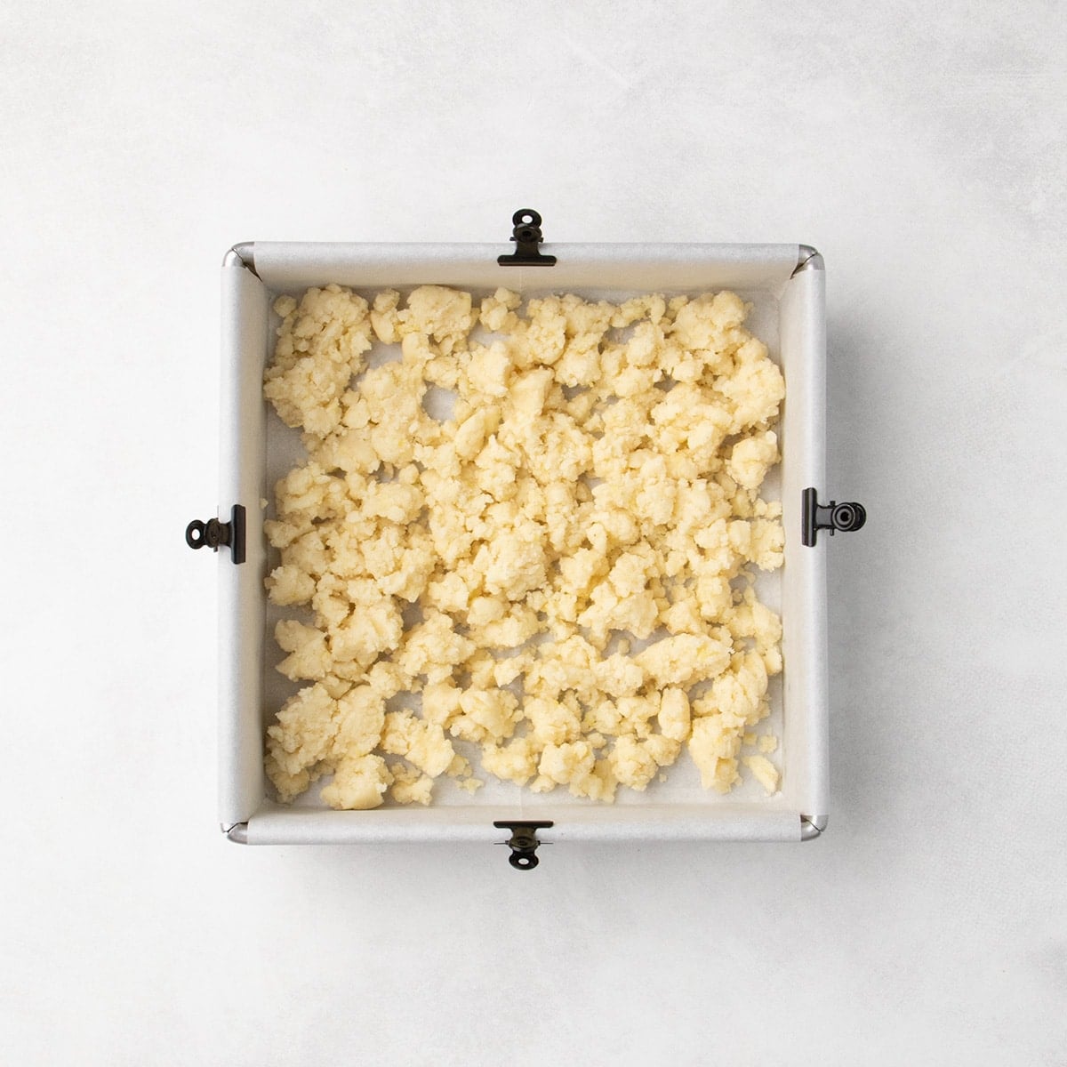 Pale dough clumps scattered in an even layer in a square baking pan lined with parchment paper, clipped to each of the four sides with a small black binder clip.