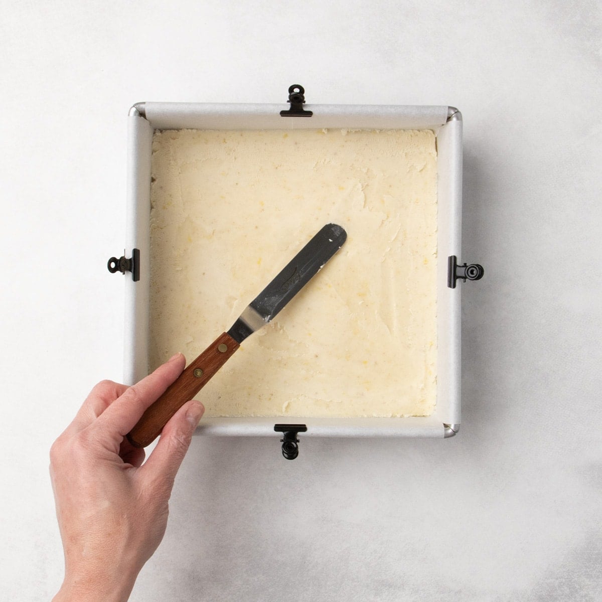 Hand holding a small offset spatula to press pale dough into a smooth, even layer in a parchment-lined square pan.