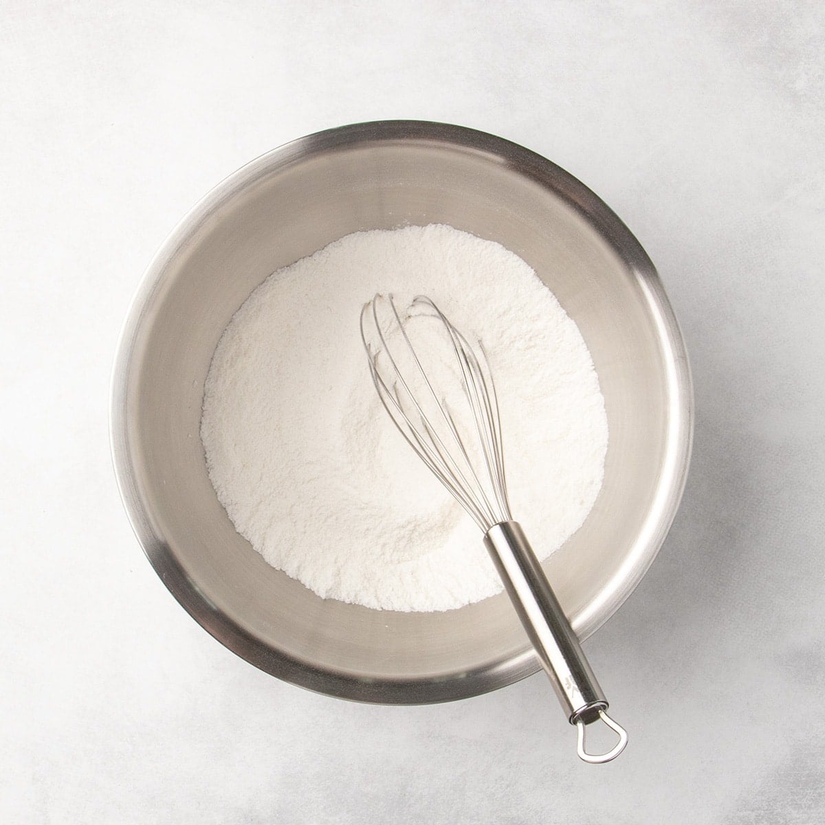 Metal mixing bowl with white dry ingredients and a whisk resting inside.