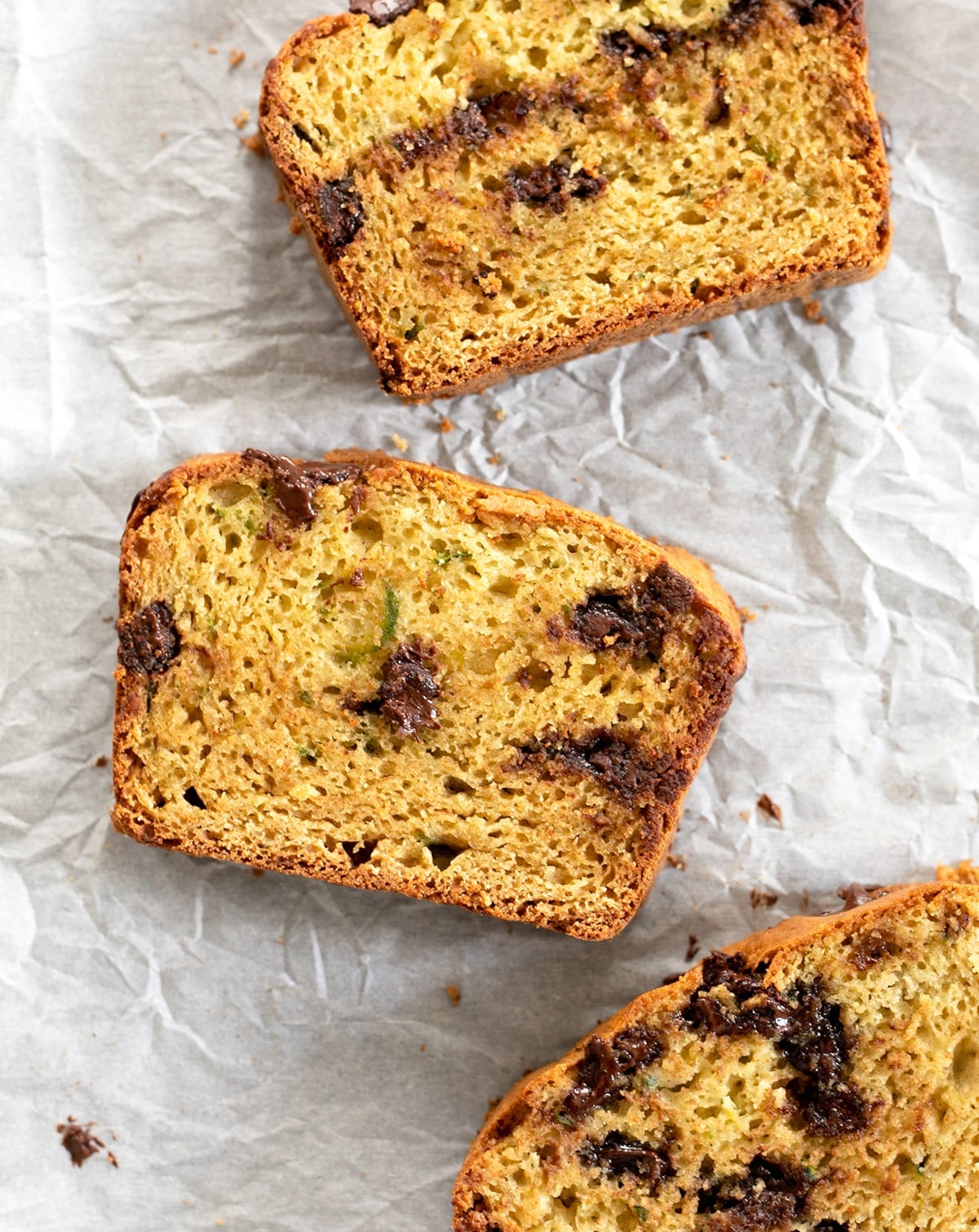 Overhead image of 3 slices the bread with green flecks and chocolate chips lying flat on wrinkled white paper.