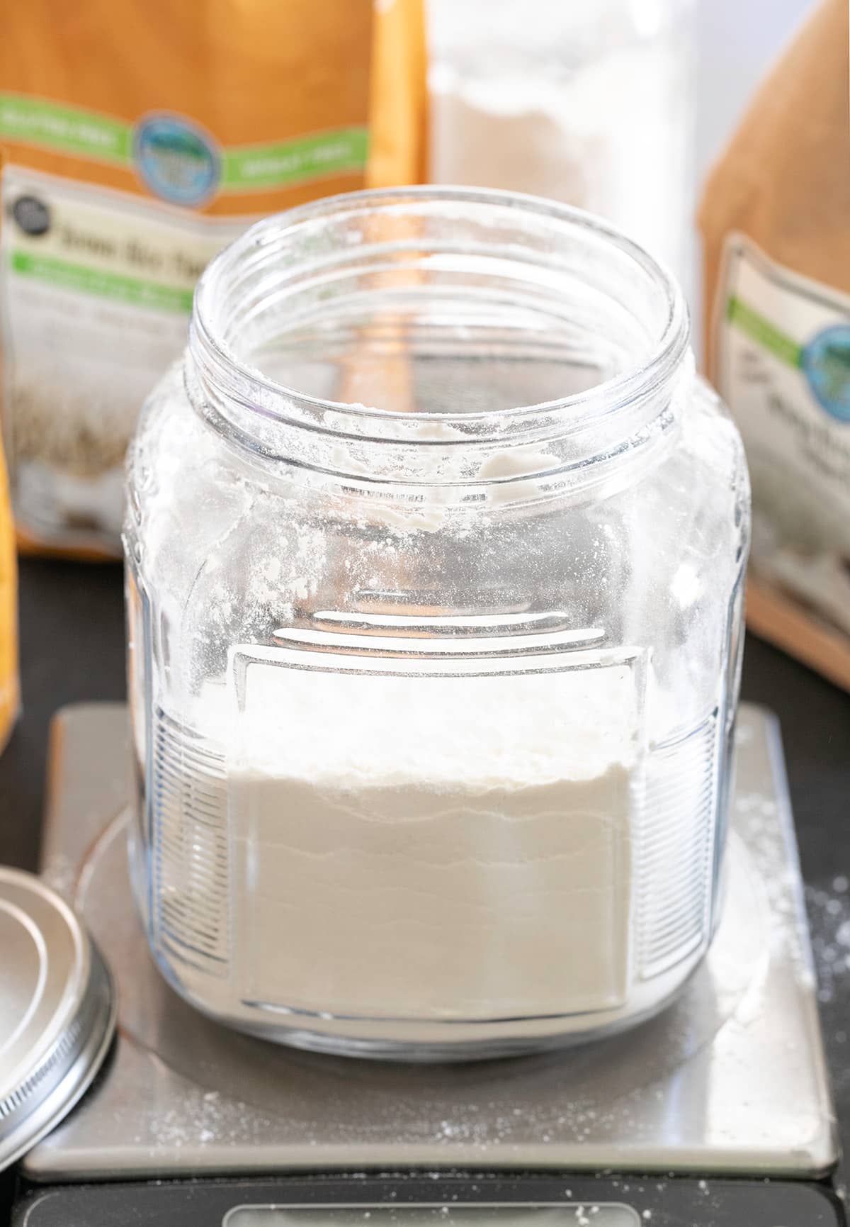 white powder gluten free flour blend in a large open jar on digital scale with the metal round lid nearby