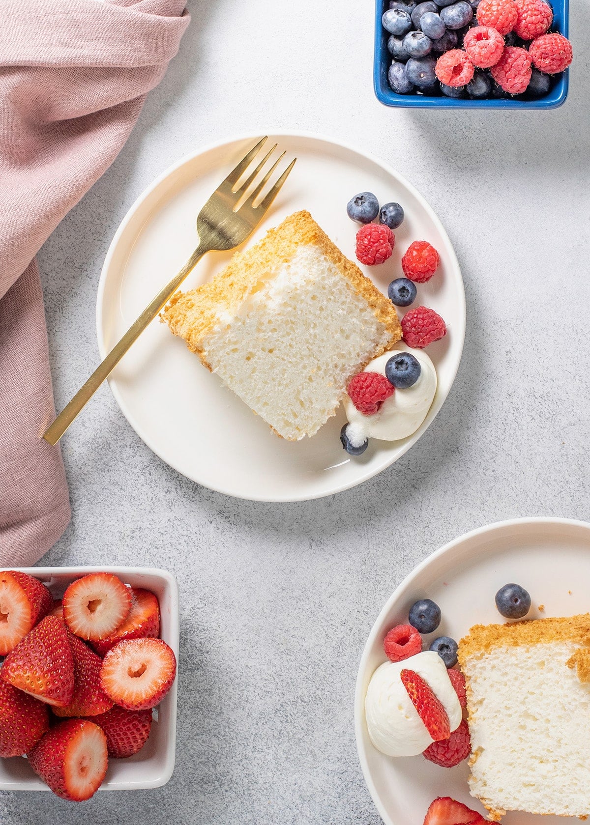Overhead image of 2 white plates with gluten free angel food cake slice berries and cream one with a fork and extra berries