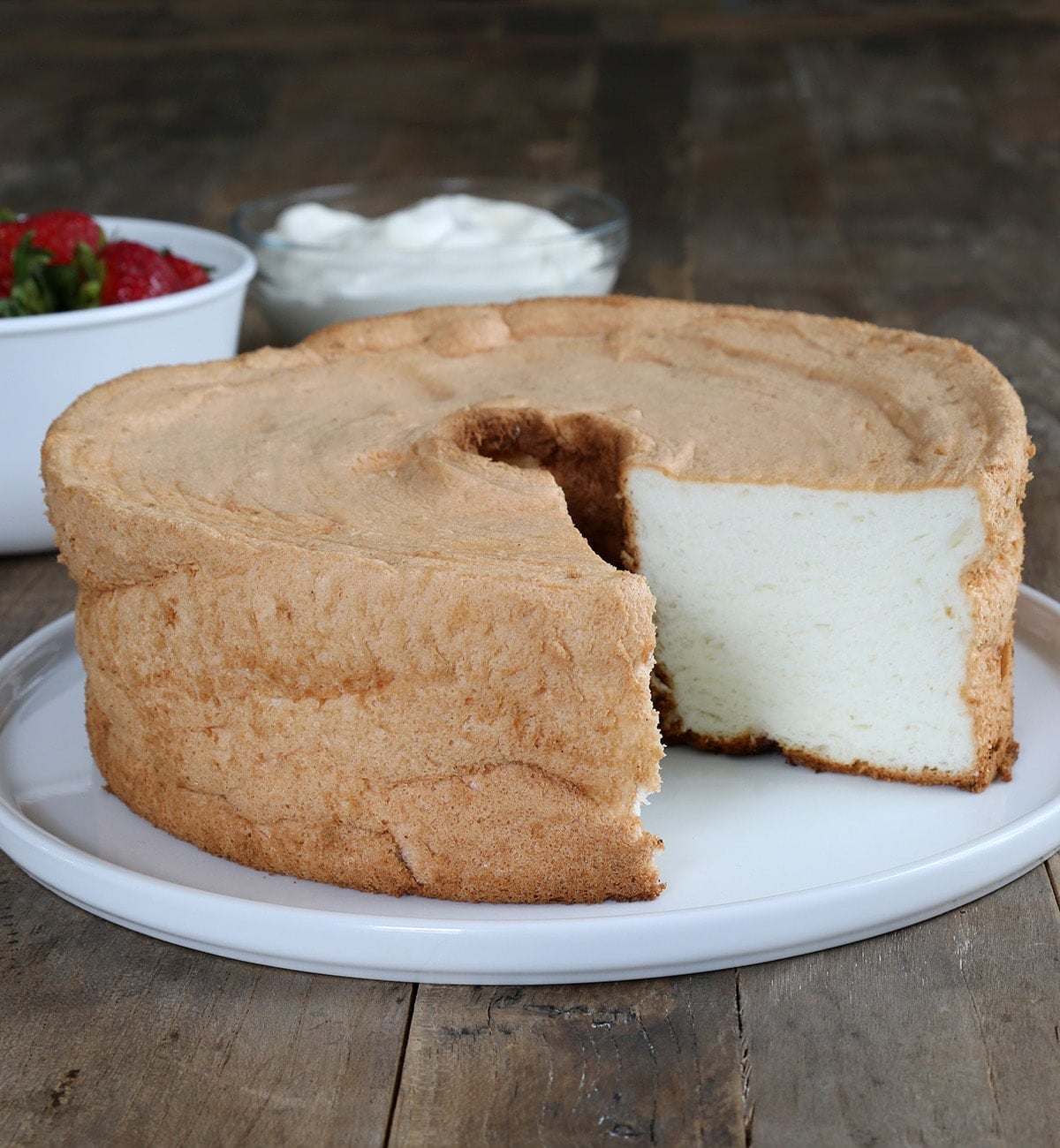 tube pan shaped gluten free angel food cake with light brown crust with slice out of it on white serving plate on brown wood surface