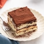 Square slice of gluten free tiramisu on a scalloped white plate with cocoa powder on top and a fork beside it.