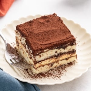Square slice of gluten free tiramisu on a scalloped white plate with cocoa powder on top and a fork beside it.